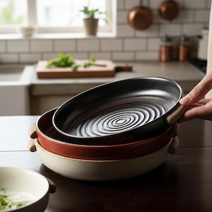 Puede incluir: Una pila de tres fuentes de cerámica en un entorno de cocina. La fuente superior es negra, la del medio es roja y la inferior es de color crema. Cada fuente tiene un asa de madera y un diseño interior acanalado.