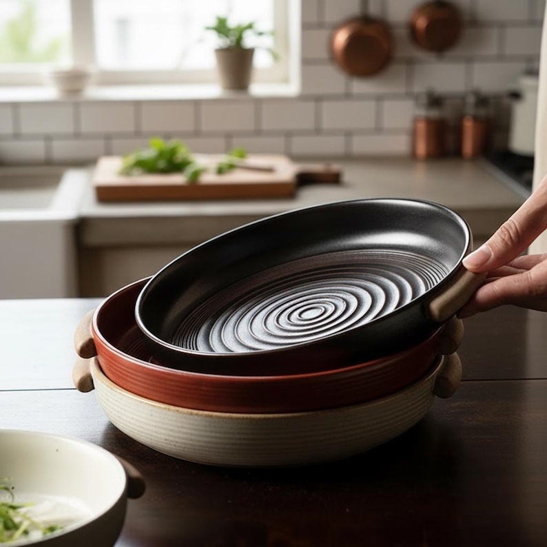 May include: A stack of three ceramic serving dishes in a kitchen setting. The top dish is black, the middle is red, and the bottom is cream-colored. Each dish has a wooden handle and a ridged interior design.