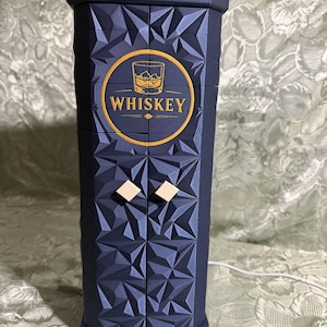 May include: A navy blue whiskey cabinet with a geometric design. The cabinet features a gold-colored circular emblem with the word "WHISKEY" and a graphic of a whiskey glass. Two small, square, cream-colored knobs are on the front.