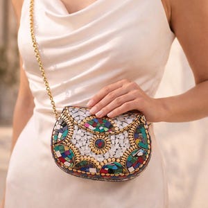 Può includere: Pochette a mosaico con tracolla a catena dorata. La pochette presenta uno sfondo bianco con forme geometriche colorate e dettagli dorati. La borsa è tenuta da una persona che indossa un vestito bianco.
