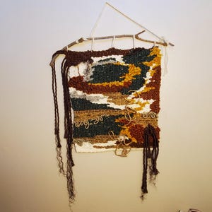 Op de afbeelding: Een geweven wandkleed met een natuurlijk kleurenpalet. Het textielkunstwerk heeft een mix van bruine, groene, witte en gele tinten, met getextureerde details en hangende franjes. Het is opgehangen aan een houten tak.