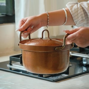 May include: A copper-colored cooking pot with a lid and two handles. The pot is sitting on a black stovetop. The pot has a hammered texture and a shiny finish. The lid has a handle on top. The pot is being held by a person.