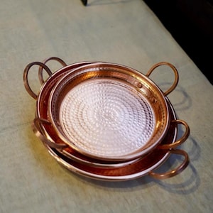 May include: A stack of three copper pans with hammered interiors and loop handles. The pans are different sizes, nested together. The copper has a warm, reddish-brown color, and the interiors have a textured, reflective surface.