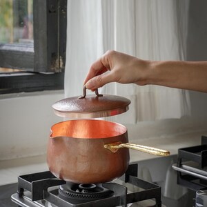 Op de afbeelding: Een koperen steelpan met een opgetild deksel. De pan heeft een goudkleurige handgreep en staat op een gasfornuis. De binnenkant van de pan is helder en reflecterend. Diameter ongeveer 15 cm.