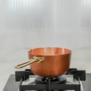 May include: A copper saucepan with a brass handle sits on a black gas stove burner. The hammered copper pot has a rounded shape and a shiny finish. The handle is attached with brass rivets.