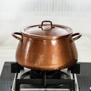 May include: A round, hammered copper pot with a matching lid and two side handles. The pot features a polished, metallic finish with visible indentations from the hammering process. It is positioned on a black stovetop burner with a metal grate.
