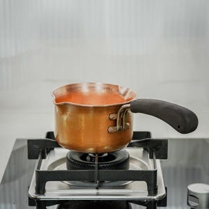 May include: A copper saucepan with a black handle sits on a gas stove. The pan contains a reddish liquid, possibly soup or sauce. The stove has a black glass top and a silver burner.