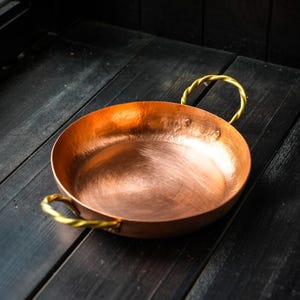 Könnte beinhalten: Eine Kupferpfanne mit zwei goldfarbenen, gedrehten Griffen. Die Pfanne hat eine breite, flache Schüssel und eine polierte Oberfläche. Die Griffe sind an den Seiten befestigt. Die Pfanne steht auf einer dunklen Holzoberfläche.