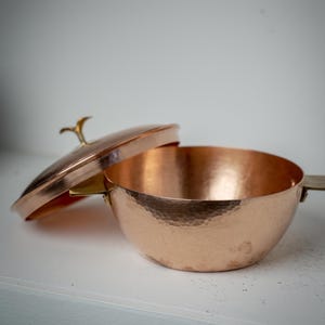 May include: A copper saucepan with a lid, handle, and hammered texture. The lid is slightly ajar, revealing the interior. The handle and lid finial are gold-coloured. The pan is on a white surface.