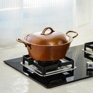 May include: A copper-colored cooking pot with a lid and two side handles. The pot is sitting on a black stovetop with a silver burner. The pot has a hammered texture and a curved handle on the lid.