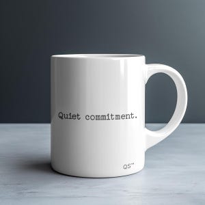 May include: White ceramic mug with a curved handle. The mug features the phrase "Quiet commitment." printed in black, centered on the front. The letters "QS™" are printed in black near the bottom. The mug is on a light gray surface.