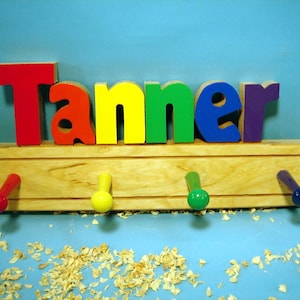 May include: A wooden coat rack with the name "Tanner" spelled out in colorful letters. The letters are red, yellow, green, blue, and purple. There are four hooks on the rack, painted red, yellow, green, and blue.