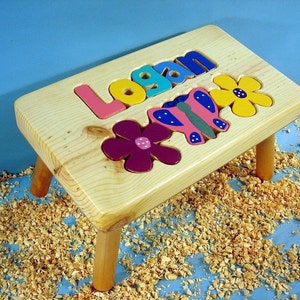 May include: A wooden step stool with the name "Logan" spelled out in colorful letters. The stool has three legs and is decorated with a butterfly and two flowers.