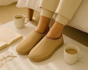 May include: A pair of tan slip-on slippers with a brown sole and a red stitched trim. The slippers are worn indoors with cream-colored pants. A white mug of coffee and an open book are nearby.