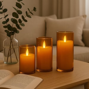 May include: Three amber glass candle holders with LED candles. The cylindrical candles emit a warm, glowing light. The holders vary in height, creating a layered effect. The candles are on a wooden table, with a vase of greenery and an open book.