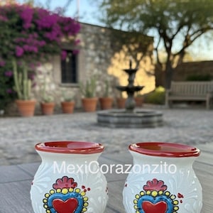 May include: Two white ceramic vases with a red rim and a heart design. The heart is red with a blue border and yellow dots. The vases have the text "MexiCorazoncito" on them.