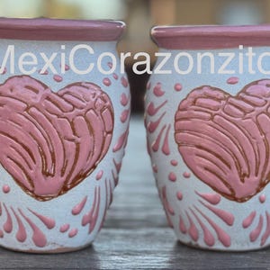 Op de afbeelding: Twee keramische vazen met een lichtblauwe basis, met een roze hartontwerp en decoratieve roze accenten. De tekst "MexiCorazonzito" is op de vazen gedrukt. De vazen hebben een roze rand.
