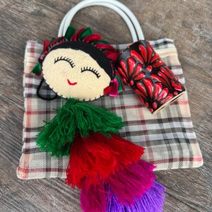 May include: A handmade tote bag with a white handle, plaid pattern, and a decorative doll face. The bag features a red and black ceramic cup and colorful tassels in green, red, pink, and purple.