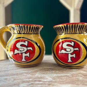 Könnte beinhalten: Zwei gelbe Keramikbecher mit dem Logo der San Francisco 49ers. Die Becher haben ein rot-weißes Logo auf einem schwarzen Oval, mit einem goldenen Griff und Rand. Der Rand hat ein schwarz-goldenes Streifendesign.