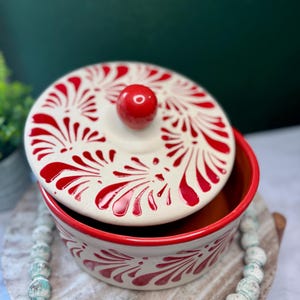 May include: A ceramic bowl with a lid, featuring a cream-colored base and vibrant red floral patterns. The rim and a small knob on the lid are also red. The bowl is likely used for serving or storage.