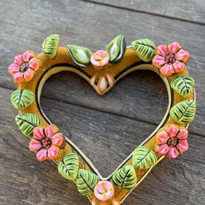 Puede incluir: Un marco decorativo en forma de corazón con un borde amarillo, adornado con flores rosas y naranjas y hojas verdes. El marco está hecho de cerámica o un material similar y tiene una apariencia rústica y artesanal. Las flores y hojas son tridimensionales.