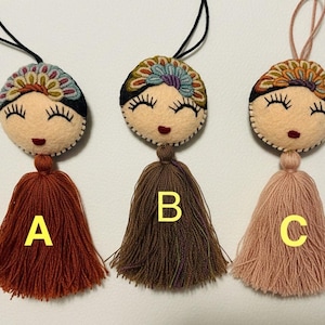 May include: Three handmade felt doll ornaments with smiling faces, long eyelashes, and colorful headbands. Each doll has a different colored tassel: rust, brown, and peach. Black string loops are attached for hanging.