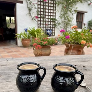May include: Two black ceramic coffee mugs with a leaf design sit on a wooden table. The mugs are filled with coffee. The background features a white building with plants and flowers in terracotta pots, creating a rustic, outdoor setting.