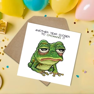 May include: A humorous greeting card featuring a cartoon frog with a weary expression. The card reads "ANOTHER YEAR CLOSER TO CROAKING IT". The image is set against a yellow background with balloons, confetti, and a brown envelope.