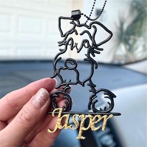 May include: Black metal dog-shaped car charm with the name "Jasper" in gold lettering. The charm is suspended from a black beaded chain. The dog design is an artistic outline.