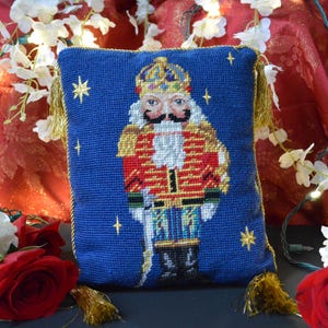 Puede incluir: Un cojín azul con una imagen bordada detallada de un soldado Cascanueces con uniforme rojo y dorado. El cojín tiene borlas doradas y está rodeado de rosas rojas y flores blancas.