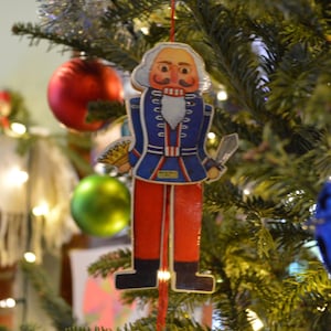 Puede incluir: Un adorno de cascanueces rojo y azul con barba blanca y una espada colgando de un árbol de Navidad.