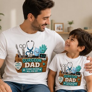 Könnte beinhalten: Zwei weiße T-Shirts mit medizinischem Thema. Die Shirts zeigen ein Holzbox-Design mit medizinischen Werkzeugen, einem Stethoskop und dem Text "SURGEON DAD" und "THANK YOU FOR ALL YOU DO". Die Shirts sind für den Vatertag.