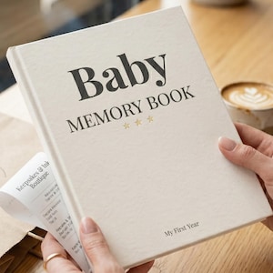 May include: A cream-colored baby memory book with the words "Baby Memory Book" in dark letters. The book has gold stars and the text "My First Year". A cup of coffee sits in the background.