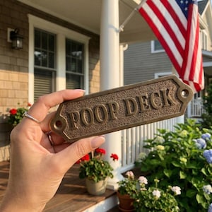 Könnte beinhalten: Ein rechteckiges, bronzefarbenes Metallschild mit strukturierter Oberfläche und erhabener Schrift mit der Aufschrift POOP DECK. Das Schild verfügt über einen dekorativen Rand und zwei kreisförmige Befestigungslöcher an den Enden.
