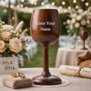 May include: A dark brown wooden wine glass with a smooth finish and a wide base. The text "Enter Your Name" is engraved on the front of the bowl. The glass is positioned on a white lace doily next to two gold rings and a scroll labeled "Marriage Vows".