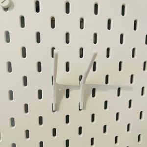 May include: Close-up of a white pegboard with evenly spaced oval holes. Two white hooks are inserted into the board. The pegboard has a smooth, matte finish. The hooks are designed to hold items.