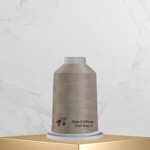 May include: A spool of Glide thread in Cool Grey 3, with a white top and base. The label includes the text "Glide 5,500 yds - Cool Grey 3" and a logo. The thread is displayed on a gold shelf against a marble background.