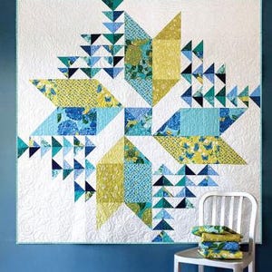 May include: A large, geometric quilt with a starburst design. The quilt features triangles and squares in shades of blue, teal, and yellow against a white background. The quilt is hanging on a blue wall. A stack of folded fabric sits on a white chair.