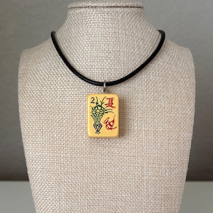 May include: A cream-colored pendant necklace featuring a rectangular tile with Chinese characters and illustrations of a vase and fish in red and green. The pendant is suspended from a black cord necklace.