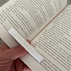 Peut inclure: Un signet blanc avec le texte en relief "Hello Beautiful!" est inséré dans un livre. Le signet est rectangulaire et fait d'un matériau souple, avec le texte vertical.