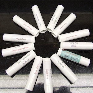 May include: A collection of 12 white lip balm tubes with various flavors, including Blue Raspberry Stushie, Pina Colada, Acai Berry, Black Cherry, Green Apple Blast, Peppermint & Honey, Vanilla Bean, Juicy Orange, Coconut Cream, Banana Coconut, Grape, and Goalie Gros.