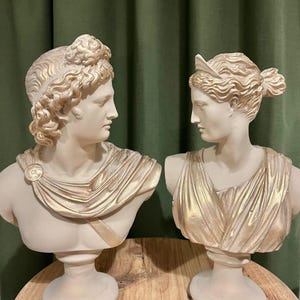May include: Two classical busts, one male and one female, face each other. The busts are off-white with gold accents on the hair and drapery. The male bust has curly hair and a draped garment. The female bust has a bun and a draped garment.