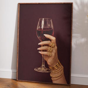 Peut inclure: Impression encadr&eacute;e d'une main tenant un verre de vin rouge. La main est orn&eacute;e de bagues et de bracelets dor&eacute;s. Le fond est bordeaux fonc&eacute; et le cadre est marron.