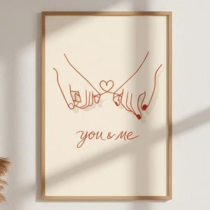Puede incluir: Un cuadro enmarcado con un dibujo lineal minimalista de dos manos entrelazadas formando un coraz&oacute;n. La obra de arte es en rojo sobre un fondo beige, con las palabras "you & me" escritas debajo.