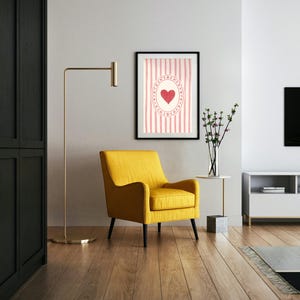 Op de afbeelding: Een moderne woonkamer met een gele fauteuil, een ingelijst kunstwerk met een hartontwerp en een gouden vloerlamp. Het kunstwerk heeft een rood hart op een roze gestreepte achtergrond. De kamer heeft houten vloeren.