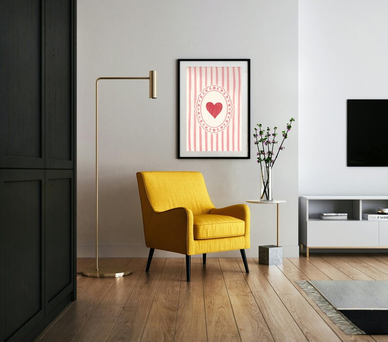 Op de afbeelding: Een moderne woonkamer met een gele fauteuil, een ingelijst kunstwerk met een hartontwerp en een gouden vloerlamp. Het kunstwerk heeft een rood hart op een roze gestreepte achtergrond. De kamer heeft houten vloeren.