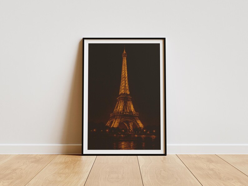 May include: Framed print of the Eiffel Tower at night. The iconic Parisian landmark is illuminated in warm tones against a dark background. The image is in a black frame, resting on a wooden floor.