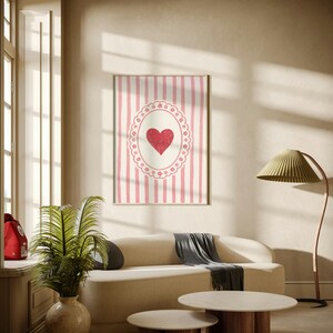 Op de afbeelding: Een ingelijste wandkunstprint met een rood hart in een wit en roze ovaal ontwerp tegen een gestreepte achtergrond. Het kunstwerk hangt boven een beige bank met een grijze plaid, aangevuld met twee ronde salontafels met houten bases.
