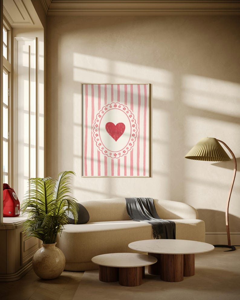 Op de afbeelding: Een ingelijste wandkunstprint met een rood hart in een wit en roze ovaal ontwerp tegen een gestreepte achtergrond. Het kunstwerk hangt boven een beige bank met een grijze plaid, aangevuld met twee ronde salontafels met houten bases.