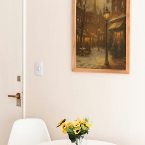 K&ouml;nnte beinhalten: Ein gerahmtes Gem&auml;lde einer verschneiten Stra&szlig;enszene mit einer Stra&szlig;enlaterne h&auml;ngt an einer Wand. Darunter steht ein kleiner wei&szlig;er Tisch mit einer klaren Glasvase mit Sonnenblumen. Ein wei&szlig;er Stuhl steht neben dem Tisch.